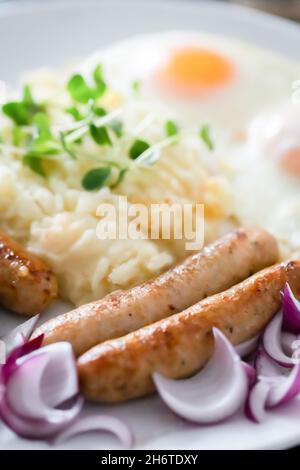 Schweinswürste mit Zwiebeln, Eiern, Kartoffelpüree und Microgreens. Herzhaftes Frühstück. Dunkler Holzhintergrund, weiße Keramikplatte. Draufsicht. Stockfoto