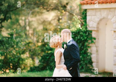 Bräutigam in einem schwarzen Anzug küsst die Braut in einem weißen Kleid vor dem Hintergrund eines grünen Parks und steinerner Tore Stockfoto
