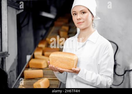 Porträt einer Bäckerin, die frisches Brot in den Händen hält, auf dem Hintergrund des automatischen Förderers der Lebensmittelbäckerei. Stockfoto