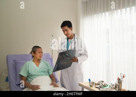 Ein asiatischer Arzt hält einen Röntgenfilm des Gehirns, in dem CT den Patienten scannt und die Ergebnisse analysiert, während er medizinische Probleme mit dem Patienten diskutiert. Stockfoto