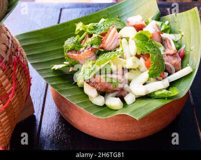 Nam Tok Moo, traditionelle thailändische Küche mit gegrillter Schweineschulter, Limettensaft, Minzblättern, Gurke, Knoblauch und Chili auf einem Palmblatt. Stockfoto