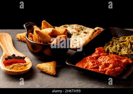 Nahaufnahme eines Tisches mit köstlichen Gerichten aus Indien. Dunkler Hintergrund mit Naan, Hühnchen, Palak Paneer, Samosa, verschiedenen Gewürzen a Stockfoto