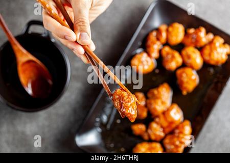 Eine Person isst köstliches Mandarinenhuhn, ein chinesisch-amerikanisches Gericht mit Holzstäbchen. Die Sauce wird in einer Porzellanschüssel serviert. Dunkles Bac Stockfoto