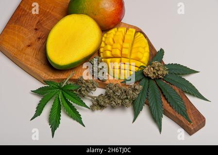Draufsicht auf geschnittene Mangofrüchte mit Cannabisblättern und Knospen. Myrcen und Terpene in Mangos unterstützen die Wirkungen von THC in Cannabis. Auf Holzschneidplatte Stockfoto