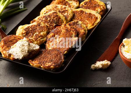 Zucchini-Pfannkuchen im türkischen Stil oder Krapfen, bekannt als Schleim, serviert auf schwarzem Teller mit frischem Gemüse und saurer Creme. Nahaufnahme des Bildes auf der Rückseite des dunklen Steins Stockfoto