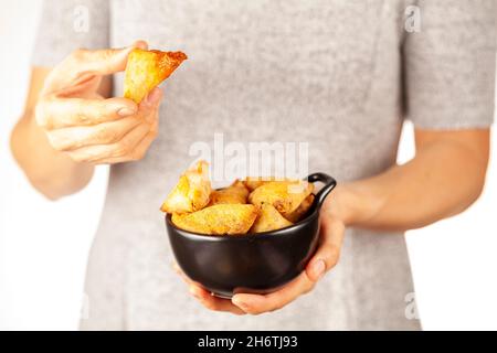 Eine Frau hält eine Porzellanschale mit indischen Samosas, dreieckigen knusprigen frittierten Snacks mit würzigen, herzhaften Füllungen. Sie isst einen. Licht und Stockfoto