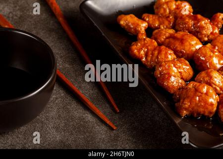 mandarine-Hühnchen in Orange auf schwarzen Porzellantellern serviert. Spezielle herzhafte Sauce wird mit Löffel hinzugefügt. Essstäbchen sind in der Nähe zu sehen. Ein chinesisches Gericht popul Stockfoto