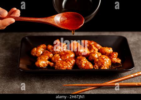 mandarine-Hühnchen in Orange auf schwarzen Porzellantellern serviert. Spezielle herzhafte Sauce wird mit Löffel hinzugefügt. Essstäbchen sind in der Nähe zu sehen. Ein chinesisches Gericht popul Stockfoto