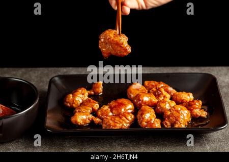 Eine Person isst köstliches Mandarinenhuhn, ein chinesisch-amerikanisches Gericht mit Holzstäbchen. Die Sauce wird in einer Porzellanschüssel serviert. Dunkles Bac Stockfoto