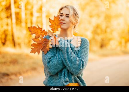 Porträt einer jungen, glücklichen Frau, die hoffentlich in die Ferne blickt, gekleidet in einen blauen Strickpullover, die Herbstblätter vor dem Hintergrund eines Stockfoto