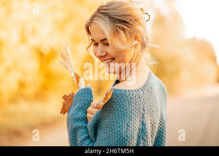 Junge, schöne Frau, blond, mit zurückgezogenen Haaren, trägt einen blauen Pullover, lächelt vor dem Hintergrund gelber Blätter und hält Herbstblätter fest Stockfoto