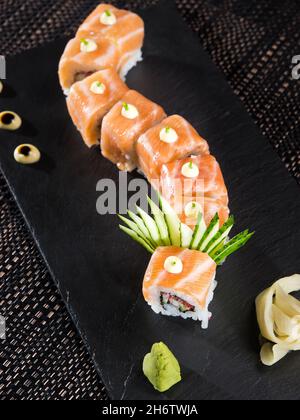 Frische Lachs-Sushi-Rollen mit Sahnetropfen, serviert mit Gurkenfächer und Wasabi auf einem Schieferteller Stockfoto