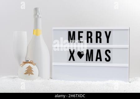 Weiße Champagnerflasche, Sektglas und Leuchtkasten mit Text FROHE WEIHNACHTEN auf einem Tisch mit Schnee. Minimales Weihnachts- oder Neujahrskonzept. Stockfoto