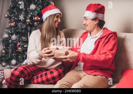 Romantisches Paar tauscht Geschenke in der Nähe geschmückten weihnachtsbaum. Mann gibt weihnachtsgeschenk Überraschung lächelnde Frau im Pullover beim Entspannen auf Stockfoto