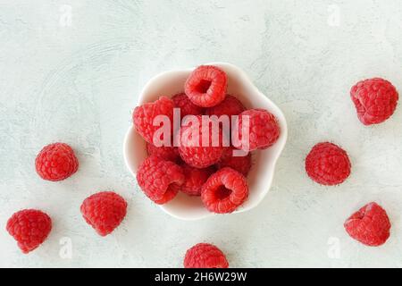 Nahaufnahme von oben mit Himbeeren in einer niedlichen weißen Schüssel und Himbeeren auf einem pastellfarbenen Texturhintergrund mit Kopierfläche. Stockfoto