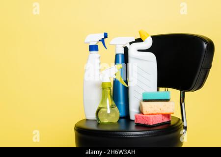 Flaschen mit Waschmittel und sauberen Schwämmen auf schwarzem Stuhl isoliert auf gelb Stockfoto