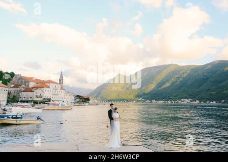 Der Bräutigam umarmt die Braut von hinten, während er auf dem Pier vor dem Hintergrund der Bucht, der Berge und der antiken Gebäude steht Stockfoto