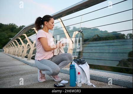 Eine ruhige, athletische Frau mittleren Alters in pinkem T-Shirt und Sport-Leggings sitzt auf einer Brücke und wickelt die Kleidung aus, hält ein Frottee-weißes Handtuch und bereitet sich auf den Morgen vor Stockfoto