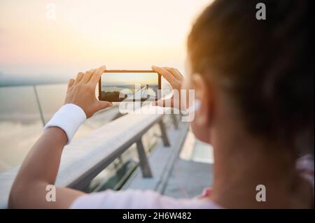 Rückansicht der Joggerin in pinkem T-Shirt mit Smartphone und Foto des wunderschönen Sonnenaufgangs am Ende der Brücke vor dem morgendlichen Joggen Stockfoto