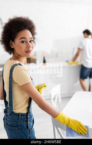 Lockige afroamerikanische Frau mit Sprühflasche und Lappen in der Nähe des Stuhls und verschwommener Mann in der Küche Stockfoto