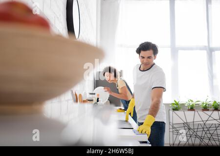 Junger Mann, der Küchenschrank mit Lappen in der Nähe von lockigen afroamerikanischen Frau in der Küche putzt Stockfoto