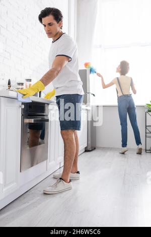 Junger Mann putzt Küchenschrank mit Lappen in der Nähe von lockigen afroamerikanischen Frau mit Staubbürste in der Küche Stockfoto