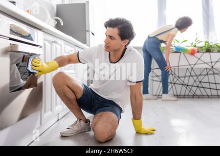 Junger Mann, der in der Küche einen elektrischen Ofen mit einem Lappen in der Nähe einer lockigen afroamerikanischen Frau mit einer Staubbürste putzt Stockfoto