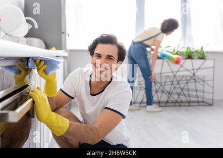 Glücklicher junger Mann, der in der Küche einen elektrischen Ofen mit einem Lappen in der Nähe einer lockigen afroamerikanischen Frau mit einer Staubbürste putzt Stockfoto