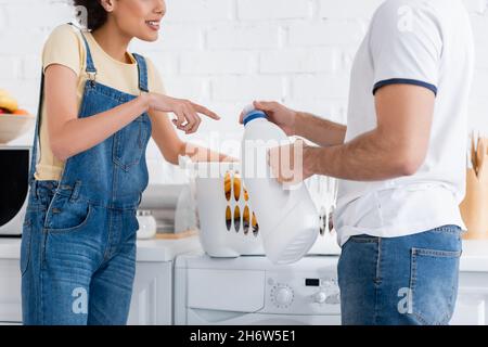 Beschnittene Ansicht einer afroamerikanischen Frau, die auf eine Flasche mit Waschmittel in der Nähe des Freundes und einen Korb mit schmutziger Wäsche zeigt Stockfoto