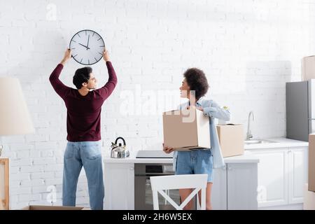 Glücklicher Mann hängende Wanduhr in der Nähe afroamerikanische Freundin mit Karton Stockfoto