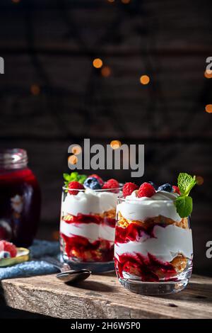 Auf einem hölzernen Hintergrund mit Lichtern, Dessert mit Rahm, Quark, Himbeeren und Heidelbeeren in einem Glas Stockfoto