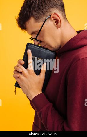 Der Christ betet mit geschlossenen Augen mit einer Bibel in seinen Händen. Der Mensch betet auf der Heiligen Bibel, die Hände gefaltet im Gebet auf der Heiligen Bibel auf einer gelben Straße Stockfoto