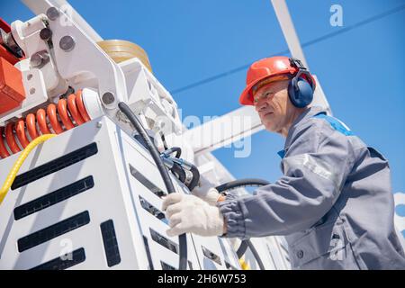 Industriearbeiter Ingenieur Mann in Schutzhelm und Kopfhörer überprüft Logistikkran im Hafen. Stockfoto