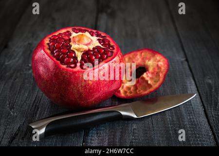 Reifer Granatapfel auf dunklem Grund und ein Holztisch mit abgeschnittener Oberseite und einem Metallmesser. Stockfoto