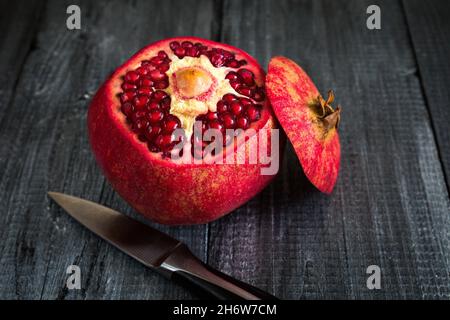 Reifer Granatapfel auf dunklem Grund und ein Holztisch mit abgeschnittener Oberseite und einem Metallmesser. Stockfoto