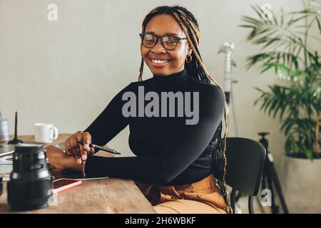 Kreative junge Frau lächelt in ihrem Heimbüro an der Kamera. Glückliche Fotografin, die einen Stylus-Stift in der Hand hält, während sie an ihrem Schreibtisch sitzt. Junger Freelan Stockfoto