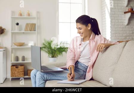 Glückliche junge Frau mit Laptop schreibt Informationen in einem Notebook, das zu Hause auf dem Sofa sitzt. Stockfoto