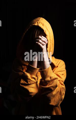 Nicht erkennbare Frau mit einer Kapuze, die auf ihren Kopf gesetzt wurde und auf einem schwarzen Hintergrund betet. Vorderansicht. Stockfoto