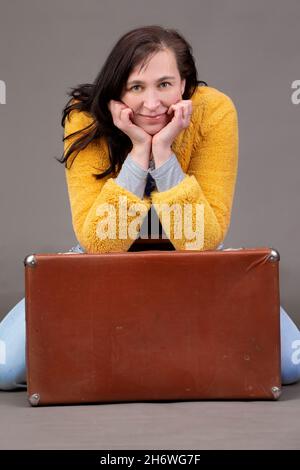Glückliche 50-jährige Frau in gelben Kleidern mit Vintage-Koffer auf grauem Hintergrund. Porträt einer Frau mittleren Alters. Stockfoto