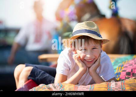 Ein lustiger kleiner Junge in einem Strohhut liegt auf einem Wagen und schaut auf die Kamera und lächelt. Stockfoto