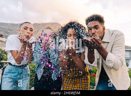 Eine Gruppe von Freunden bläst bunte Konfetti im Freien. Fröhliche junge Leute, die während einer Party auf dem Dach feiern. Multikulturelle Freunde haben Spaß t Stockfoto