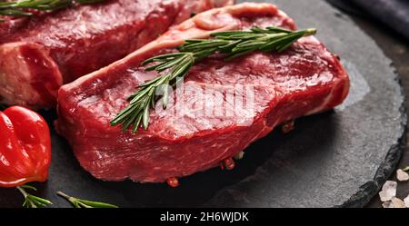 Zwei rohe Steak New York mit Rosmarin und Gewürzen auf einem Stück Pergamentpapier auf altem dunklen Steinhintergrund. Draufsicht. Modell. Stockfoto