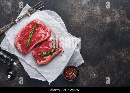 Zwei rohe Steak New York mit Rosmarin und Gewürzen auf einem Stück Pergamentpapier auf altem dunklen Steinhintergrund. Draufsicht. Modell. Stockfoto