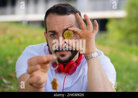 Junger Mann posiert mit neuem Hausschlüssel und Bitcoin-Münze. Konzept für junge Investoren. Stockfoto