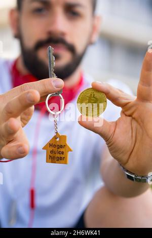 Junger Mann posiert mit neuem Hausschlüssel und Bitcoin-Münze. Konzept für junge Investoren. Stockfoto