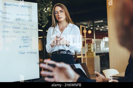 Junge Geschäftsfrau diskutiert mit ihrem Team in einem modernen Arbeitsraum über Marketingideen. Selbstbewusste junge Geschäftsführerin, die während eines Me eine Präsentation gibt Stockfoto