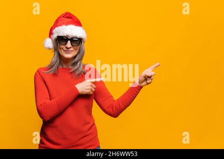 Charmante fröhliche Frau mittleren Alters in rotem weihnachtsmann Hut und stilvolle Sonnenbrille zeigt Finger weg auf leere Kopie-Raum, reifes Dame zieht die Aufmerksamkeit, isoliert auf gelb, Mock-up Stockfoto