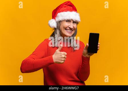 Zufriedene ältere Frau in santa hat hält Smartphone mit einem leeren Bildschirm, zeigt Daumen nach oben, fröhliche ältere grauhaarige Dame Werbung für neue mobile App, zeigt Verkauf isoliert auf gelb, Mockup-Konzept Stockfoto