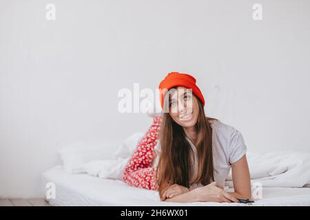 Lächelndes lustiges Mädchen in einem roten Hut und Weihnachten Pyjamas, liegt im Bett mit weißen Laken. Feiertage zu weihnachten. Gute Weihnachtsstimmung Mädchen. Stockfoto