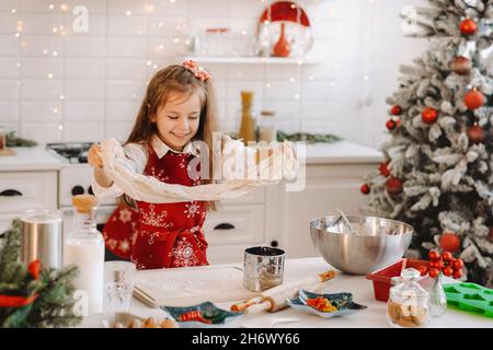 Ein kleines Mädchen in einer roten Schürze in der Neujahrsküche bereitet Teig für Weihnachtskekse zu. Stockfoto